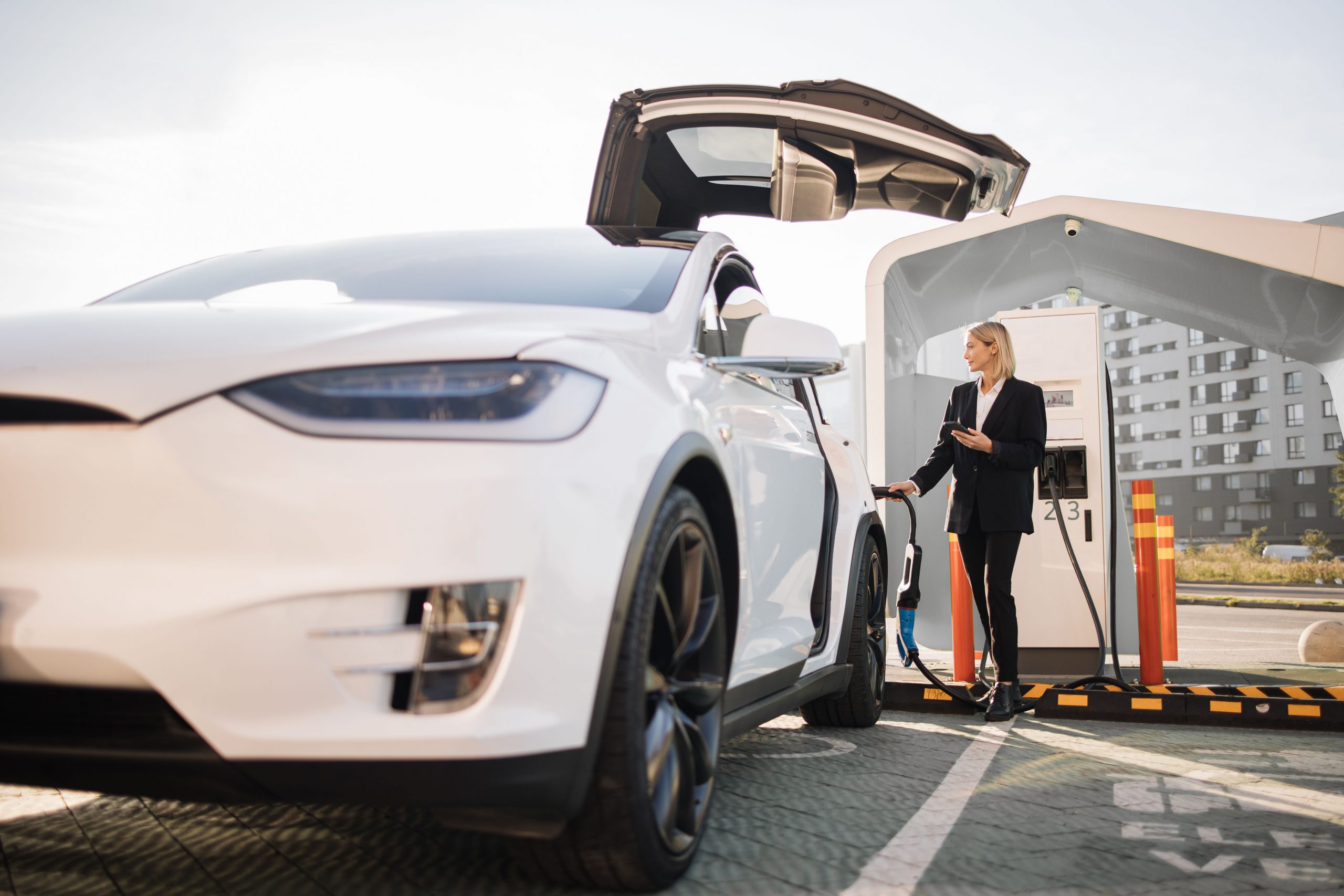 Beautiful caucasian woman in formal wear using modern smartphone while charging white lectro car at station. Concept of sustainable and economic transportation.